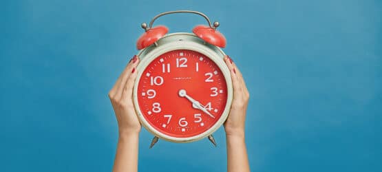 Bild von einem roten Wecker, der von zwei Händen nach oben gehalten wird