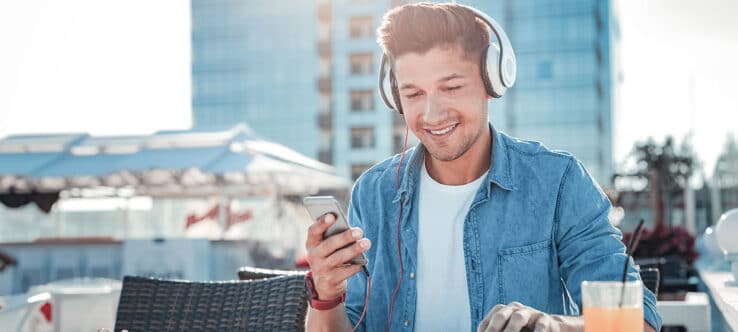 Bild von einem jungen Mann, der mit Kopfhörern und einem Smartphone auf einer Terrasse sitzt