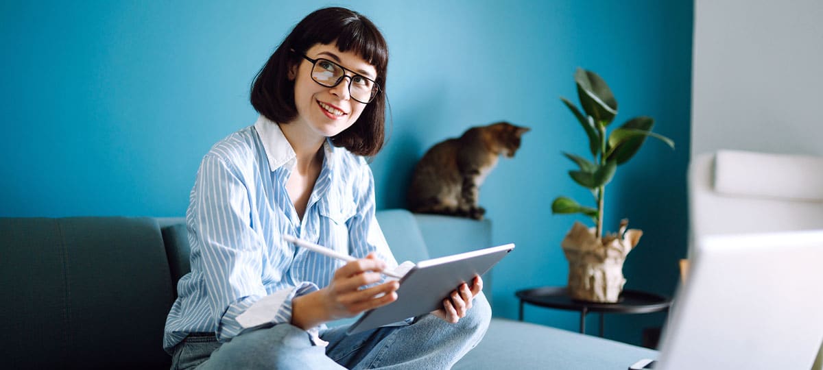 Foto von einer lächelnden Frau mit Brille, die ein Tablet in der Hand hält. Im Hintergrund sitzt eine Katze auf dem Sofa,